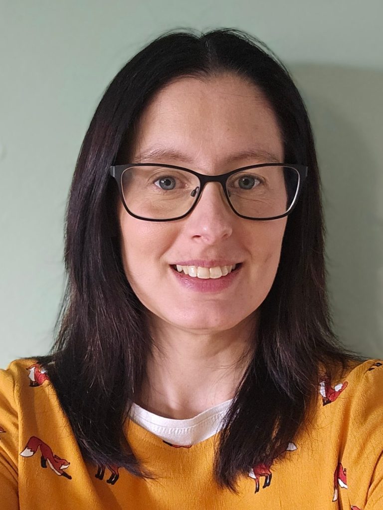 Georgina Judd Winchester Speech Therapist has mid length brown hair, green eyes, a big smile and is wearing a maroon coloured top.